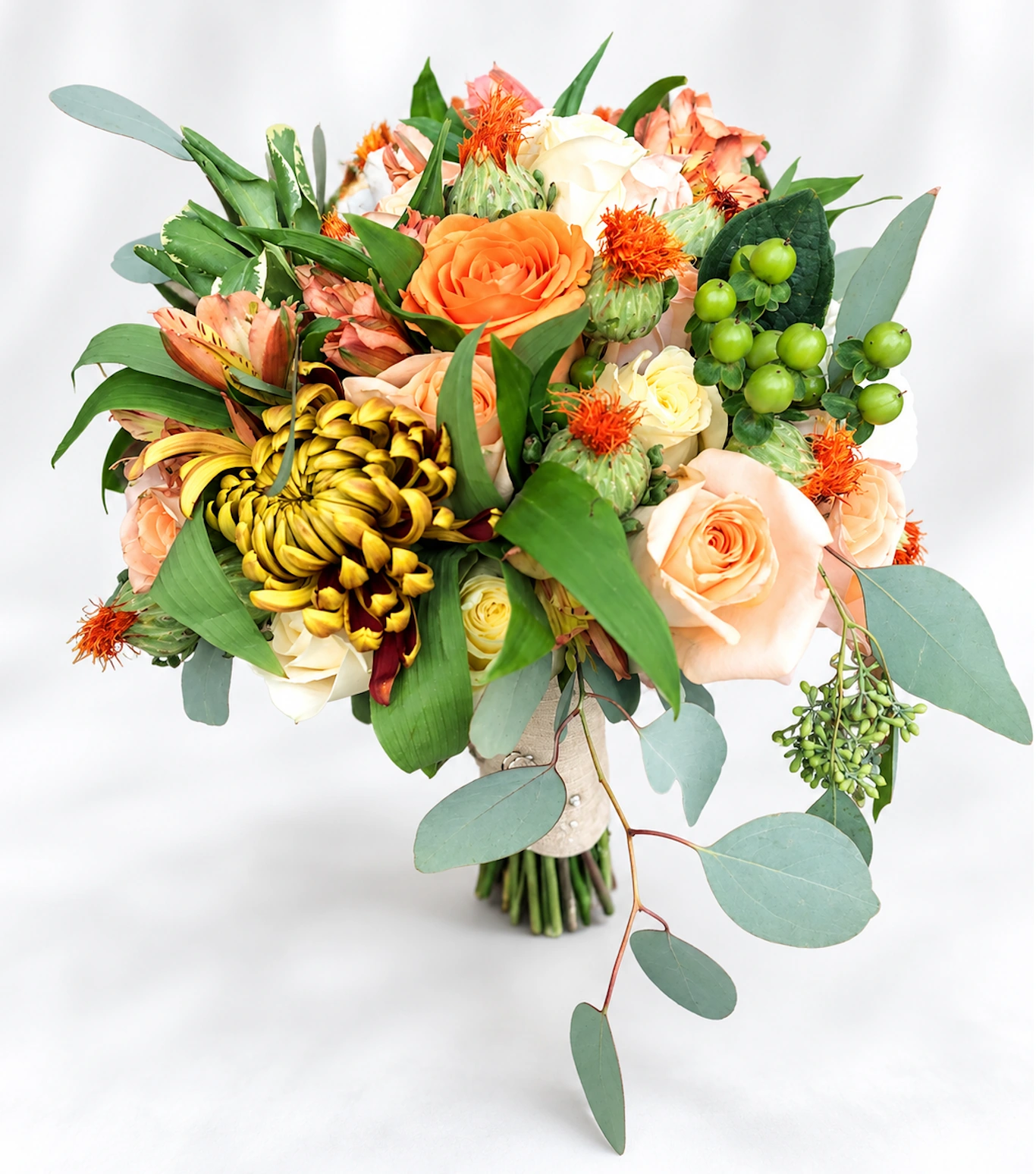 Bridal bouquet of peach, orange and ivory roses, green berries, and trailing eucalyptus against a soft white background