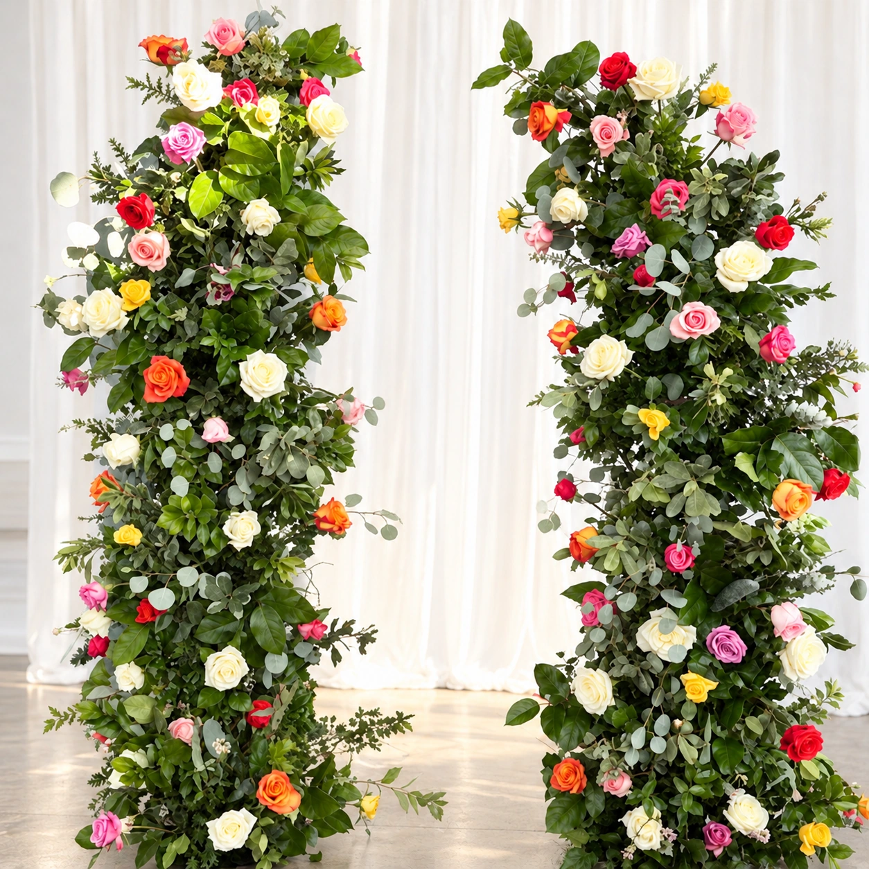 Two tall floral pillars with pink, white, red, orange and yellow roses, lush greenery and an ivory curtain backdrop